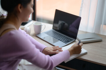 Asian female student writing on notebook while using laptop computer, studying online course at home
