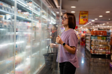 Asian customer holding shopping basket choosing frozen food from glass door refrigerator in supermarket
