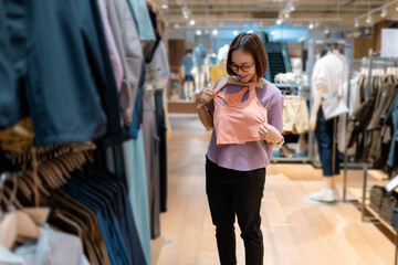 Asian woman shopping for clothes in a retail store, holding a pink top on a hanger