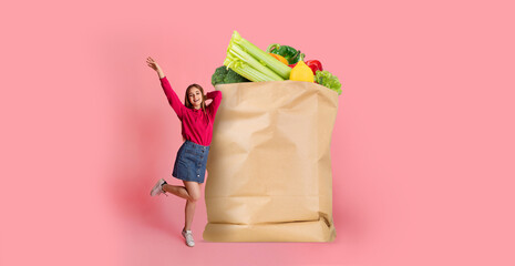 A happy shopper stands next to a large paper bag filled with fresh vegetables and fruits. The backdrop is bright pink, enhancing the upbeat atmosphere of the shopping experience.