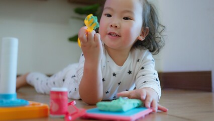 Cute Asian toddler child girl playing with modeling clay on Wooden Floor, enhancing creativity education and motor skills in a cozy home environment.