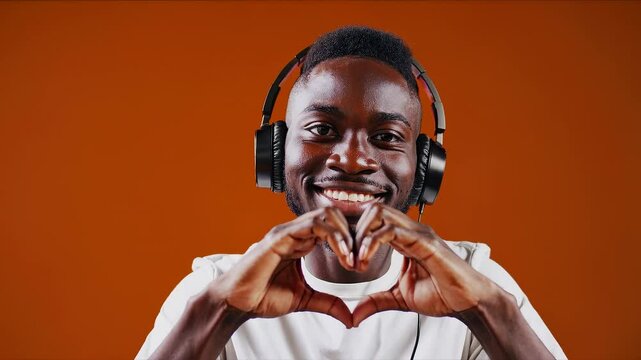 Smiling young man with headphones making heart gesture with hands in front of orange background