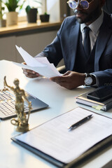 Vertical shot of African American male lawyer, wearing glasses and suit, sitting in office and studying documents