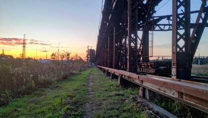 Obraz premium Industrial Sunset Over Abandoned Bridge with Grassy Pathway