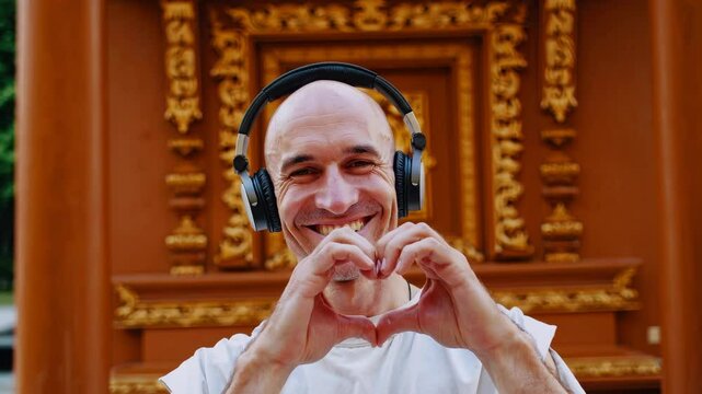 Smiling bald man with headphones making heart gesture with hands in front of ornate temple architecture