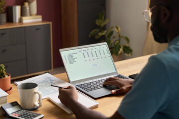 Over shoulder shot of young Black man sitting at desk at home, using laptop to plan family budget