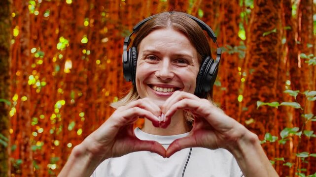 Smiling woman with headphones making heart gesture with hands in lush forest setting