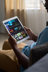 Vertical medium shot of African American man sitting on couch at home, watching movie or TV show trailer on streaming app on tablet