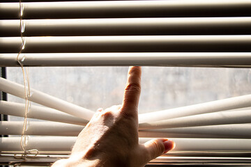 Woman's hand lifting blinds to look out the window in sunlight,hand