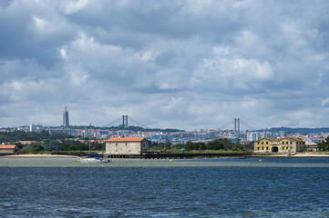 Panoramic view of Lisbon skyline from Seixal, with the Tejo River, a sailing boat, bridge, and monument under dramatic cloudy sky.