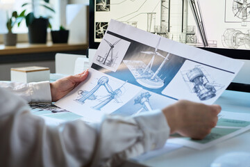 Unrecognizable engineer sitting at office desk holding printed paper with wind turbine drawings