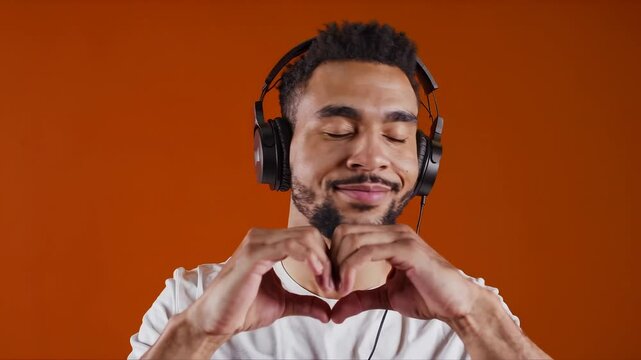 Young man enjoying music with headphones, making a heart gesture with hands on orange background