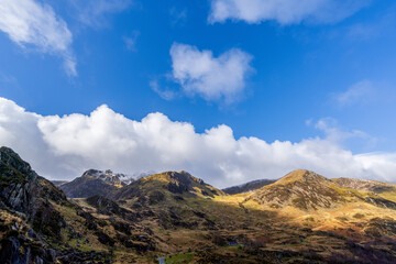 Distant view of Cwm Idwal peaks framed by billowing cumulus clouds over sunlit scree slopes and autumnal grassland