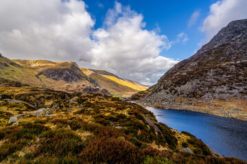 Sunlit Llyn Idwal nestled at the base of rugged scree slopes and wind-swept moorland under a dramatic bank of cumulus clouds