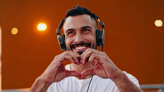 Smiling man listening to music with headphones, making heart gesture with hands outdoors at sunset