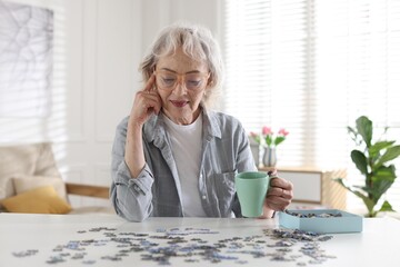 Woman with cup of drink solving jigsaw puzzle at white table indoors