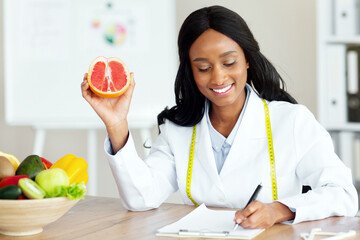 Attractive African American nutrition adviser holding grapefruit, making meal plan for client at...