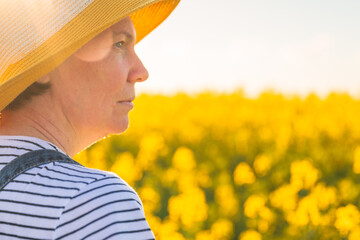 Rear view of female farmer with straw hat standing in blooming rapeseed field and looking into...