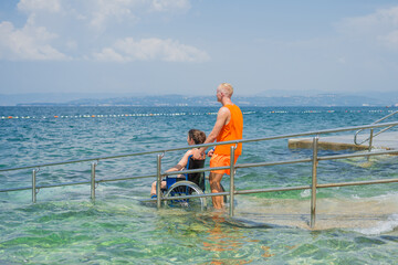 Male assistant pushing a woman with disability in a wheelchair into the wavy sea across an...