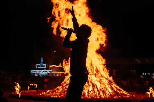 Ein S&auml;nger performt auf einer B&uuml;hne. Im Hintergrund brennt ein gro&szlig;es Lagerfeuer / Maifeuer / Hexenfeuer.