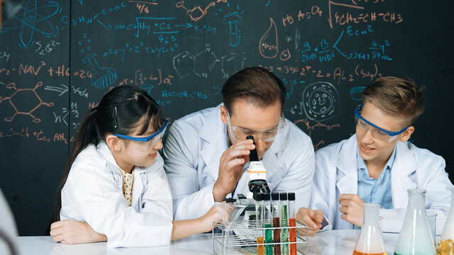 Smart teacher looking at microscope surrounded by diverse student. Happy instructor and group of happy diverse children doing experiment at blackboard with theory in STEM science class. Edification.