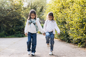 Obraz premium Two young girls walking hand in hand along a sunlit pathway surrounded by lush greenery, enjoying a joyful moment of friendship and connection