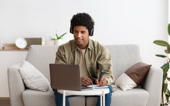 Web-based education concept. Happy teen guy in headset taking notes while communicating online with tutor on laptop. Funky African American adolescent making homework for his online course
