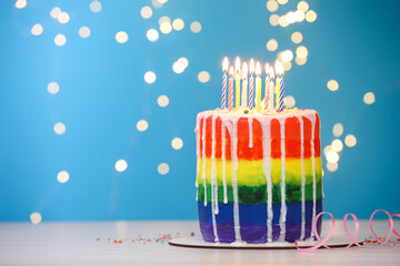 Delicious rainbow birthday cake with burning candles, sprinkles and streamer on light table against...