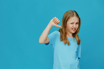 Young girl showing a thumbs down gesture against a bright blue background, expressing...