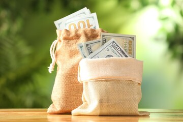 Money bags with dollar banknotes on wooden table against blurred green background, closeup