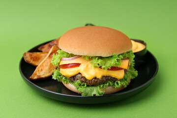 Tasty cheeseburger with sauce and potato wedges on green background, closeup