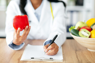 Cropped view of African American nutrition adviser holding bell pepper, planning meal for client at clinic. Weight loss specialist writing prescription for patient at medical office. Selective focus