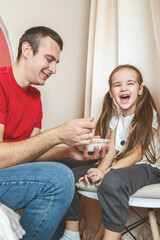 Cheerful small girl daughter and her handsome young dad spending time together at home. Father feeding his little daughter.