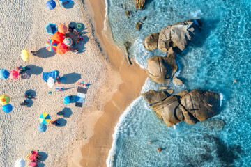 Aerial view of colorful umbrellas, sandy beach and blue sea with waves and rocks at sunset. Summer in Sardinia island, Italy, Tropical seascape. Top drone view of clear water, relaxing people, beach