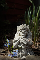 Old concrete gargoyle sitting in the sun in a shady garden with little blue flowers.