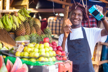 african market man selling fruits items holding a pos terminal and card looking at camera