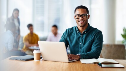 Fototapeta premium A man with short brown hair and black-framed glasses, wearing a teal blue shirt, is shown sitting at a table with a silver laptop, and a blurred background featuring coworkers engaged in conversation,