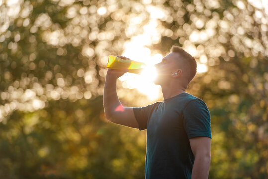 A fit young man drinking water from a bottle after a run or workout in a summer park, active healthy lifestyle.