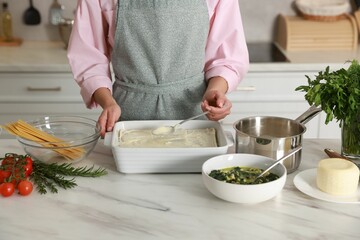 Woman spreading bechamel sauce onto spinach lasagna at marble table indoors, closeup