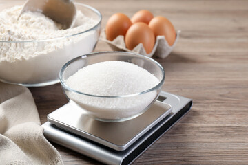 Digital kitchen scale with bowl of sugar and other products on wooden table, closeup