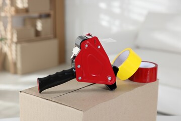 Dispenser with adhesive tape rolls on cardboard box indoors, closeup