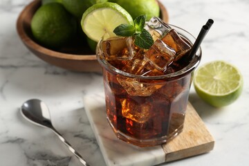 Tasty Cuba Libre cocktail in glass, limes and spoon on white marble table, closeup