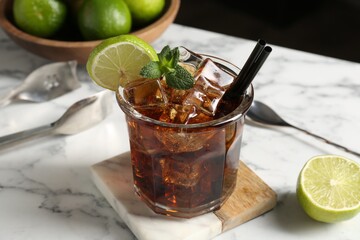 Tasty Cuba Libre cocktail in glass, limes, tongs and spoon on white marble table, closeup