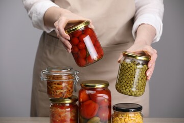Woman with jars of different pickled products at table against grey background, closeup