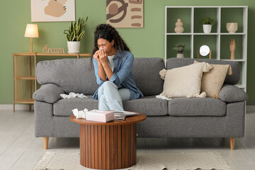 Young African-American woman sneezing in handkerchief and sitting on sofa at home. Allergy concept