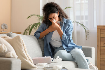 Young African-American woman sneezing in handkerchief and sitting on sofa at home. Allergy concept