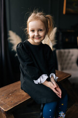 Adorable 5 years old girl in black dress top and blue tights sitting on wooden bench, hands clasped, looking at camera with gentle smile on face, creating heartwarming portrait of childhood innocence.