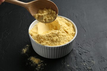 Taking corn flour with spoon from bowl on black textured table, closeup