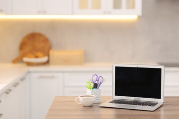 Fototapeta premium Laptop with blank screen, cup of hot drink and stationery on wooden table in kitchen. Mockup for design