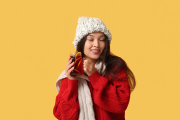 Beautiful young happy Asian woman in winter clothes with glass jar of hot mulled wine on yellow...